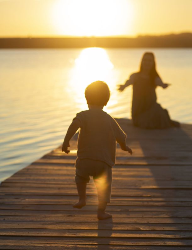 A woman stretches out her hands to encourage chlid to hug her, on a dock with the sun in the background.